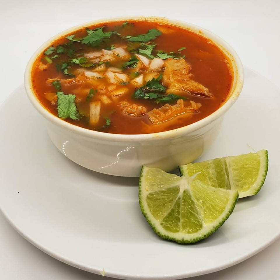 A bowl of red menudo soup with tripe, onions, and cilantro, served with lime wedges.