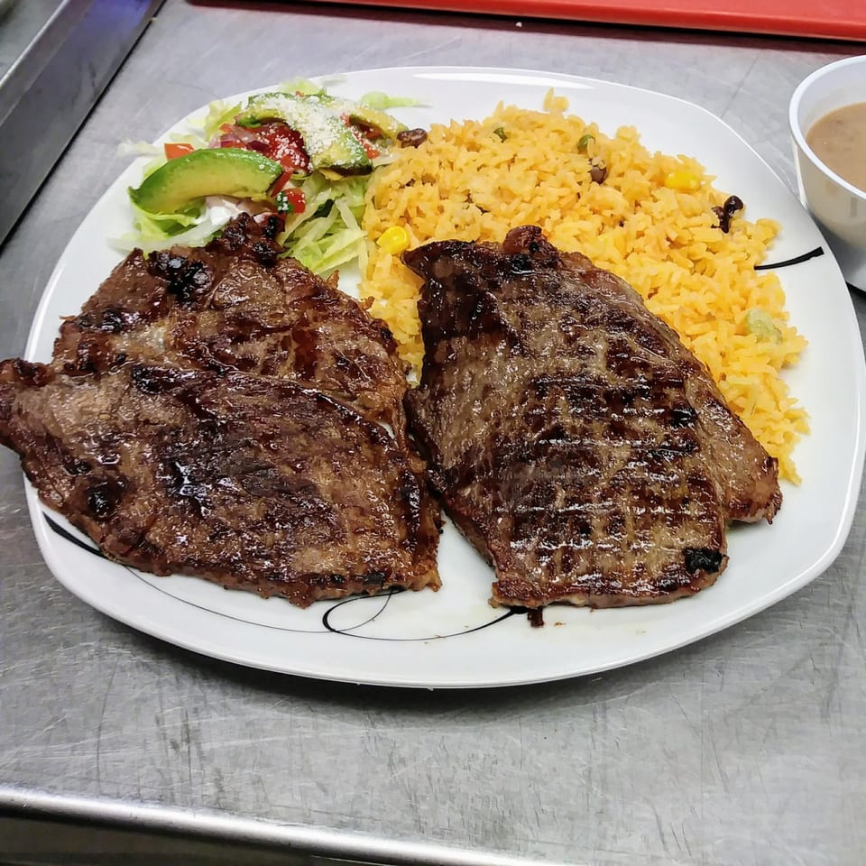 Two grilled steaks, yellow rice, and a side salad with avocado on a white plate.
