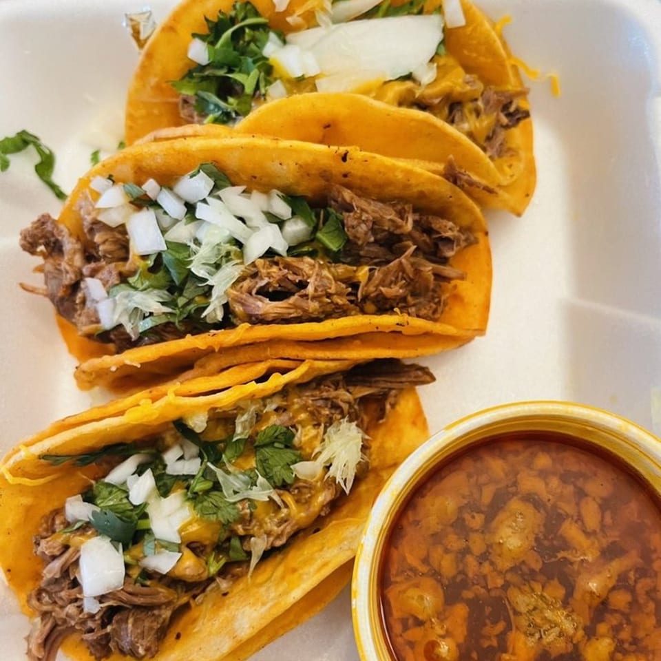 Three birria tacos with shredded beef, onions, cilantro, and a side of consommé.